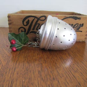 May include: A silver tea infuser with a chain, featuring a perforated, egg-shaped design. A sprig of green leaves and red berries is attached. The infuser rests on a wooden surface, with a wooden box in the background.
