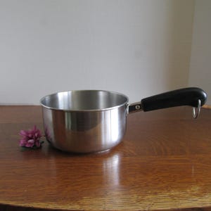 May include: A small, stainless steel saucepan with a black handle and a hanging loop. The pan is sitting on a wooden surface, with a purple flower to the left. The pan is designed for cooking and is likely used in a kitchen.