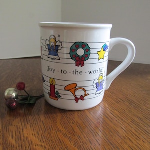 May include: White ceramic mug with a curved handle, decorated with musical notes, angels, a wreath, and a star. The mug has the text "Joy - to - the - world!" printed on it. A festive Christmas-themed mug.