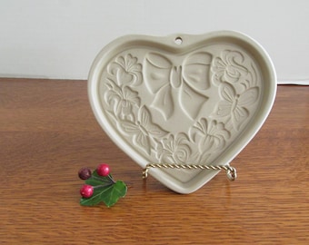 May include: A beige heart-shaped cookie cutter with a raised design of a bow and butterflies. The cutter is on a gold metal stand.