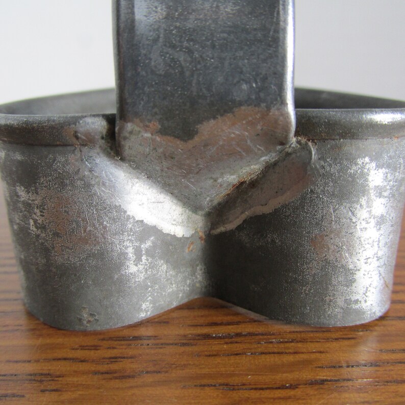 May include: A vintage metal cookie cutter in the shape of a heart with a handle. The cutter is made of silver-colored metal with some rust spots.