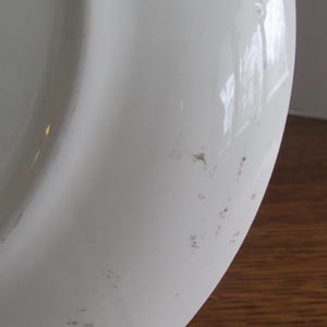 May include: White ceramic plate with a slight curve and a few small, faint purple marks on the surface.