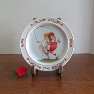 May include: A white ceramic decorative plate featuring a central illustration of two children jumping rope in a grassy field. The rim is decorated with red floral motifs and green vines. The text reads June and Sarah Stilwell Weber Calendar Collection.