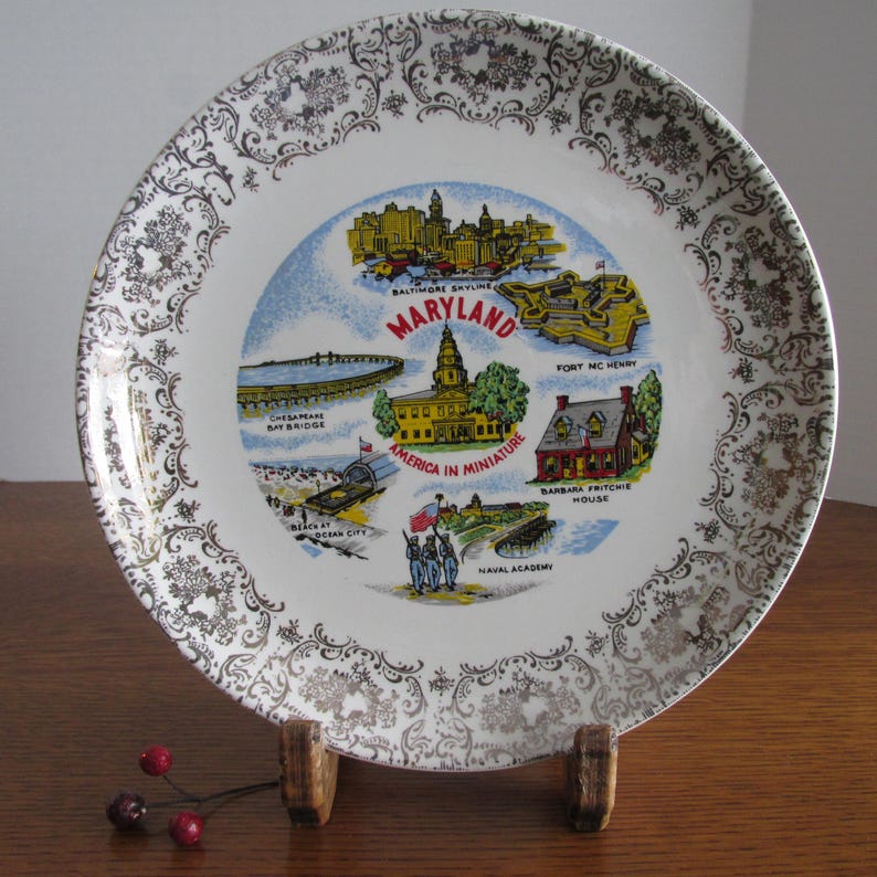 May include: A white ceramic plate with a gold rim and a decorative design. The center of the plate features a colorful illustration of landmarks in Maryland, including the Baltimore skyline, Chesapeake Bay Bridge, Fort McHenry, and the United States Naval Academy. The text "Maryland" and "America in Miniature" are printed on the plate.