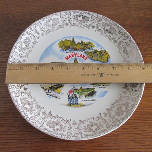 May include: A white ceramic plate with a gold rim and a decorative pattern. The plate features a map of Maryland with various landmarks, including the Baltimore skyline, Ocean City, and the Naval Academy. The text "MARYLAND" is printed in red and white.