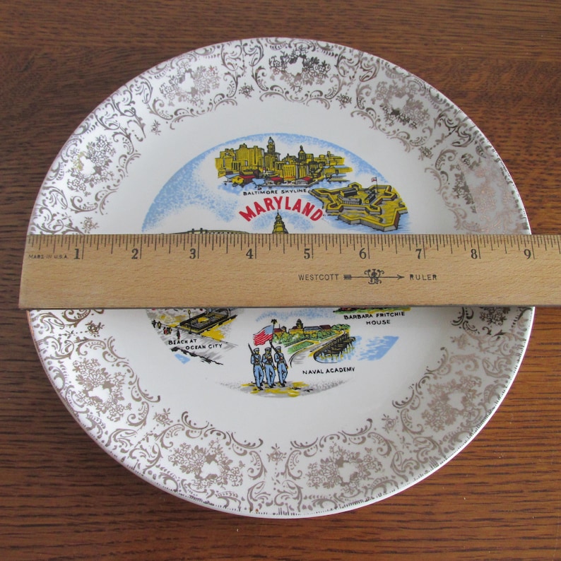 May include: A white ceramic plate with a gold rim and a decorative pattern. The plate features a map of Maryland with various landmarks, including the Baltimore skyline, Ocean City, and the Naval Academy. The text "MARYLAND" is printed in red and white.