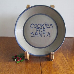 May include: A ceramic plate with a blue rim and the words "COOKIES FOR SANTA" in blue lettering. The plate is displayed on a wooden stand, with a sprig of holly and red berries in front.