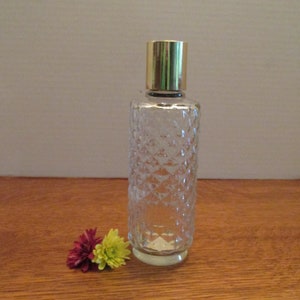 May include: A clear glass perfume bottle with a diamond pattern and a gold cap. The bottle is cylindrical and sits on a wooden surface. Two small flowers, one maroon and one yellow, are next to the bottle.