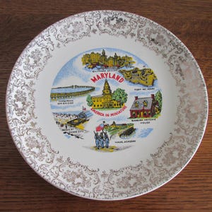 May include: A white ceramic plate with a gold rim and a decorative design. The center of the plate features a colorful illustration of various landmarks in Maryland, including the Chesapeake Bay Bridge, Fort McHenry, and the State House. The text "Maryland" and "America in Miniature" are printed on the plate.
