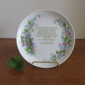 May include: A white porcelain plate with a floral design featuring blue and purple flowers. The plate has a poem printed on it that reads: "Each time you look up in the sky Or watch the fluffy clouds drift by, Or stop to pick a daffodil, Or gather violets on some hill... Or touch a leaf or see a tree. It's all GOD whispering "This is Me. And I am Faith and I am Light And in Me There shall be No Night."