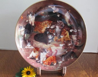 May include: Decorative plate featuring a detailed illustration of numerous kittens in various poses. The plate has a gold rim and a warm, brown background. A yellow sunflower rests in front of the plate.