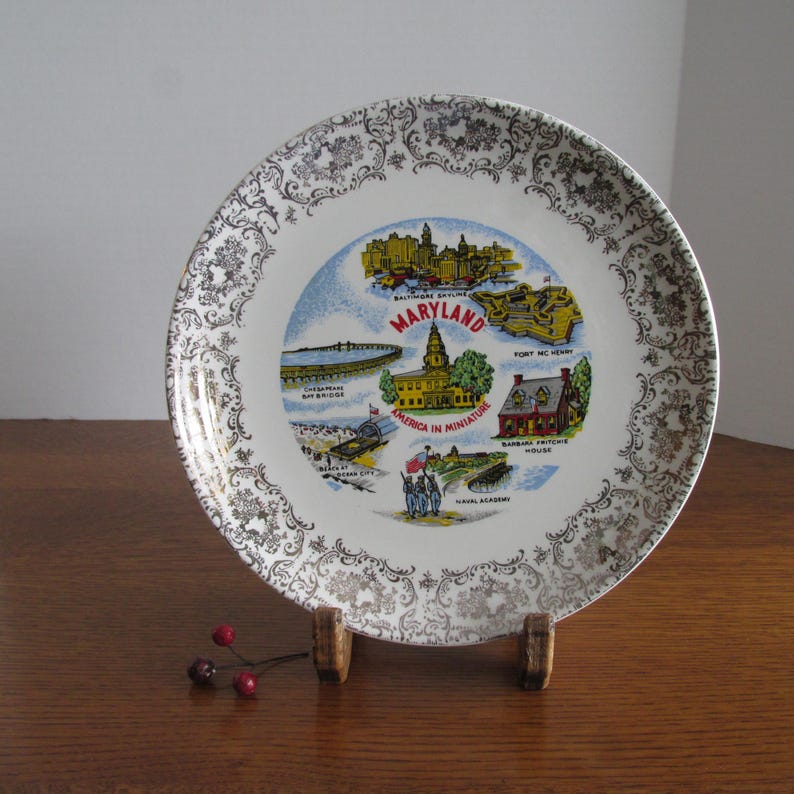 May include: A white ceramic plate with a gold rim and a decorative design. The plate features a colorful illustration of landmarks in Maryland, including the State House, Fort McHenry, and the Chesapeake Bay Bridge. The text "Maryland" and "America in Miniature" are printed on the plate.