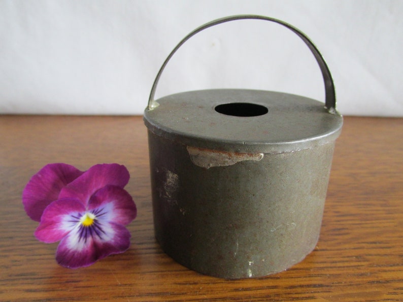 May include: A vintage metal cake mold with a handle. The mold is green and has a circular shape with a hole in the center. The mold is sitting on a wooden surface.