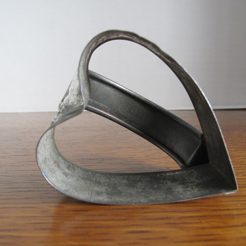May include: A vintage metal heart-shaped cookie cutter with a worn, silver finish. The cutter is slightly bent and has a rustic appearance.