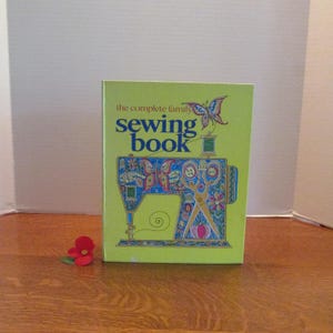 May include: A hardcover book titled The Complete Family Sewing Book with a lime green cover. The front features a blue and multicolored illustration of a sewing machine decorated with floral patterns and a butterfly, plus a small red fabric flower.