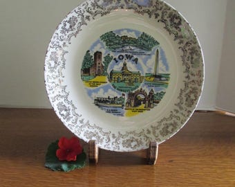 May include: A vintage decorative plate featuring the state of Iowa. The plate has a white background with a gold filigree border. The center of the plate displays various Iowa landmarks and the word "IOWA".