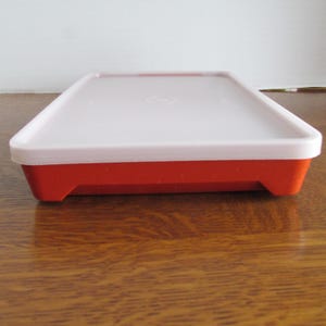 May include: A rectangular food storage container with a red base and a white lid. The lid has a slightly raised edge and a subtle design in the center. The container is made of plastic and is designed for food storage.