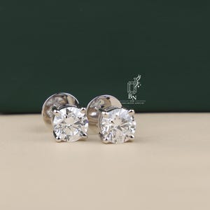 May include: A pair of round-cut stud earrings featuring clear, faceted stones set in four-prong silver-toned metal settings. The earrings include circular backings and are positioned near a logo with the text BN USA DIAMOND JEWELRY.