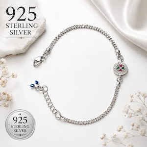 Puede incluir: Una pulsera de cadena de plata de ley 925 con un dije circular engastado con pequeñas piedras rojas y verdes rodeadas de cristales transparentes. La pulsera incluye un cierre de mosquetón y una cadena extensora con dos cuentas azules.