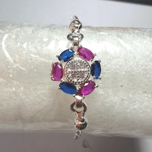 May include: A silver-toned bracelet featuring a central circular cluster of four clear stones surrounded by alternating oval-shaped blue and pink stones. The metal links connect to the central piece, which is set against a crinkled, translucent material.