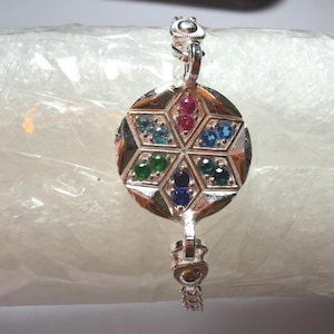 May include: A silver bracelet featuring a circular centrepiece with a geometric hexagonal pattern. Each of the six sections contains two small round gemstones in various colours, including red, light blue, dark blue, and green hues.