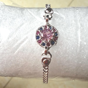 Puede incluir: Una pulsera de tono plateado con un dije circular central engastado con una piedra rosa grande rodeada de pequeñas piedras azules y rojas alternadas. La cadena consta de eslabones con detalles triangulares texturizados cerca del centro.