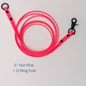 May include: A 1/2 inch hot pink leash coiled in a circular pattern. The leash features matte black hardware, including a circular O-ring at one end and a swivel trigger snap hook at the other, both secured with two black rivets on the pink strap.