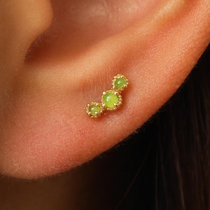 May include: A gold-toned ear stud featuring three small, round, light green stones arranged in a curved, ascending line. Each stone is set within a delicate, scalloped gold metal bezel, creating a cluster design that follows the ear cartilage.