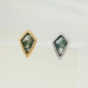 May include: Two kite-shaped stud earrings featuring moss agate stones with green inclusions. One earring is set in a gold-toned metal frame, while the other is set in a silver-toned metal frame. Both earrings have a pointed, geometric design.