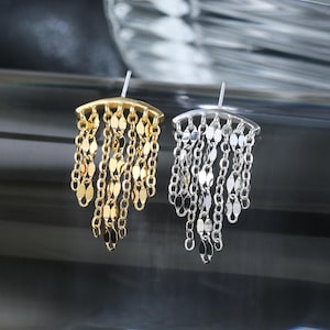 May include: A pair of stud earrings featuring a curved top bar with multiple dangling chains. One earring is gold-toned and the other is silver-toned. Each earring has several delicate chains with small, flat, reflective, oval-shaped metal links.
