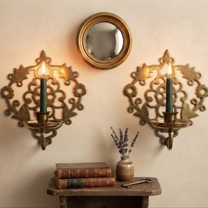 May include: Two ornate brass wall sconces with intricate cut-out patterns, each holding a dark green taper candle with a lit flame. Between the sconces is a small round mirror with a gold frame. Below are two stacked books, a vase with lavender, and a key.