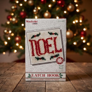 May include: A WonderArt by Caron latch hook kit box featuring a white, red, and green Noel design. The packaging displays the text Noel, 12 x 12 inches, and Quick & Easy. The box has a light gray background with a subtle snowflake pattern.