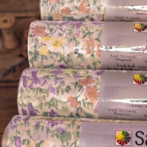 May include: A stack of five cylindrical rolls of wallpaper featuring a floral pattern with purple, yellow, and orange flowers on a light background. Each roll has a label with the text Easy to Hang and a colorful circular logo.