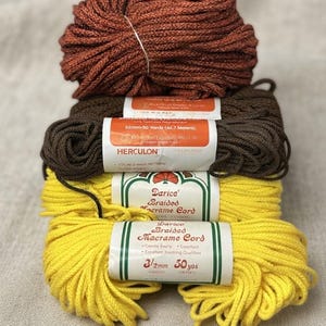 May include: Four skeins of braided macrame cord stacked vertically. The top skein is rust-colored, the second is dark brown with a Herculon label, and the bottom two are bright yellow with Darice Braided Macrame Cord labels stating 3.5mm and 50 yards.