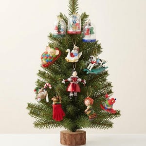 May include: A small artificial evergreen tree on a wooden base decorated with various hanging ornaments, including three glass-domed figures labeled 1992, 1993, and 1994, a mouse in a sleeping bag, a swan, a rocking horse, and several small character figurines.