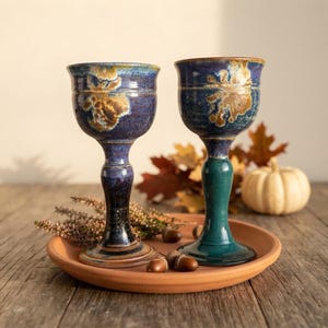 May include: Two ceramic goblets with flared rims and slender stems. The left goblet features a deep blue glaze with mottled brown accents. The right goblet has a similar blue and brown bowl atop a solid dark green stem and base.