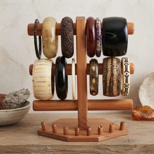 May include: A wooden jewelry display stand with two horizontal bars holding various bangles. The collection includes bracelets in black, cream, brown, and patterned designs, featuring smooth, carved, and textured surfaces arranged on the stand.