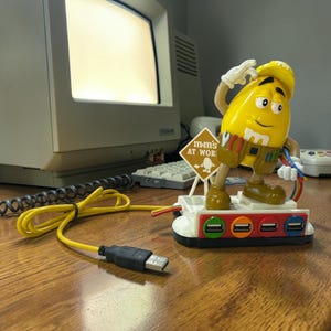 May include: A yellow M&M character figurine wearing a construction hat and tool belt, mounted on a white USB hub base with four colored ports. The base features a small sign reading M&M'S AT WORK and a yellow USB cable with a black connector.