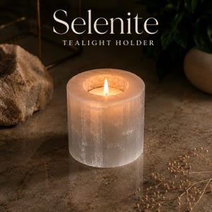 Op de afbeelding: Een cilindrische seleniet theelichthouder met een doorschijnende, witte, vezelige textuur. Een brandend waxinelichtje staat in de centrale opening en verspreidt een warme gloed. De tekst "Selenite TEALIGHT HOLDER" staat bovenaan de afbeelding.