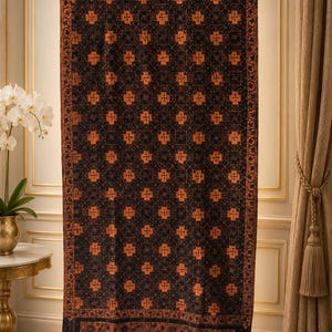 May include: A rectangular textile hanging featuring a dark black base colour with a repeating pattern of orange geometric cross-like shapes. The fabric has a wide decorative border with intricate floral motifs and a fringed bottom edge.