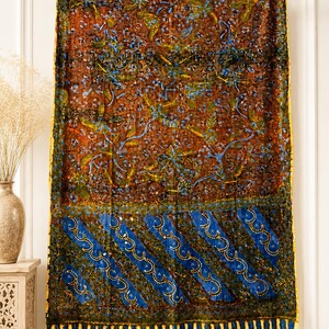 Könnte beinhalten: Ein rechteckiger Wandbehang aus Textil mit einem Muster im Batik-Stil. Der obere Bereich zeigt einen braunen Hintergrund mit blauen und grünen Blumenmotiven. Der untere Bereich hat diagonale blaue Streifen mit weißen Punkten und einen gelben Rand.