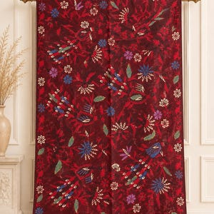 Puede incluir: Un tapiz rectangular de tela con un fondo de color burdeos intenso que presenta un patrón repetitivo de aves estilizadas y motivos florales en tonos azul, morado, verde y blanco, acabado con un borde decorativo en la parte inferior.