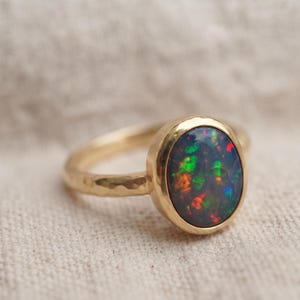 Peut inclure: Une bague en or ornée d'une opale noire de forme ovale présentant des éclats de couleur vert et rouge vifs. L'anneau de la bague possède une finition martelée texturée et est serti dans un chaton en or lisse et poli qui maintient la pierre.
