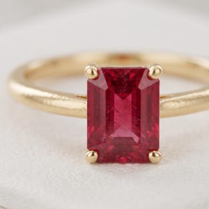 May include: A gold ring featuring a rectangular emerald-cut red gemstone set in a four-prong setting. The band is smooth and polished, showcasing a classic design with a vibrant, deep red stone as the central focal point of the jewelry piece.