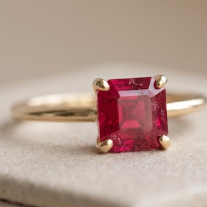 Peut inclure: Une bague en or ornée d'une pierre précieuse rouge foncé de taille carrée, maintenue par quatre griffes en métal. L'anneau est fin et poli, présentant une finition métallique lisse qui complète la pierre rouge facettée au centre du bijou.