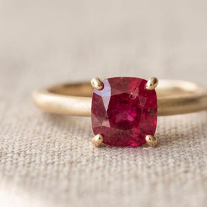 Puede incluir: Un anillo de oro con una piedra preciosa de color rojo intenso en talla cojín, montada en un engaste de cuatro garras. El aro tiene un acabado mate liso y un perfil redondeado. La piedra roja vibrante es el foco central del diseño.