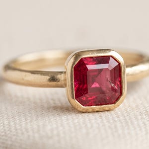 Pode incluir: Um anel de ouro com uma pedra preciosa vermelho-escura de corte quadrado, fixada numa cravação de bisel lisa. A banda do anel tem um acabamento arredondado e polido, e a pedra central apresenta uma superfície facetada que reflete a luz.