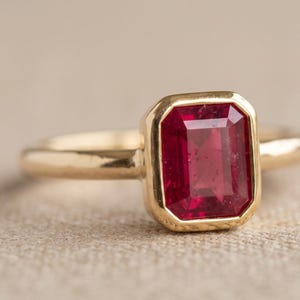 Può includere: Un anello in oro con una pietra preziosa rosso intenso di forma rettangolare a taglio smeraldo, montata in una lunetta liscia. La fascia è in oro giallo lucido con un profilo arrotondato, caratterizzata da un design pulito e lineare.