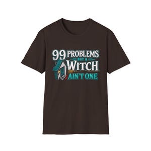 May include: A dark brown short-sleeve t-shirt featuring a graphic print with the text 99 PROBLEMS BUT A WITCH AIN'T ONE. The design includes a small blue house with a lit lantern and a brown broomstick positioned horizontally across the centre.