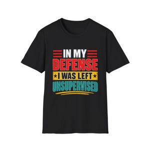 May include: A black short-sleeved t-shirt featuring a graphic print with the text IN MY DEFENSE I WAS LEFT UNSUPERVISED. The design uses red, yellow, and light blue lettering with horizontal lines and two stars, all set against a solid black background.